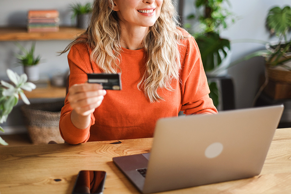 An image of a woman holding a credit card while sitting at a wooden table with a laptop