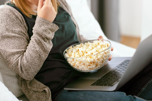 An image of a woman sitting on the couch with a laptop and a bowl of popcorn
