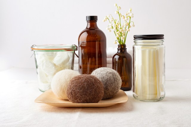 An image of dishwasher tablets in dissolvable wrappers stored in mason jar, liquid laundry soap in refillable bottle, laundry soap sheets in re-useable jar, wool dryer balls on bamboo plate, flower arrangement in re-useable bottle