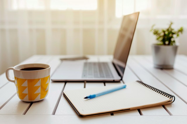 An image of a cup of coffee, a laptop, and a notebook on desk