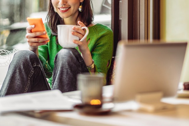 An image of a woman holding a mug of coffee and her phone at a table with a laptop