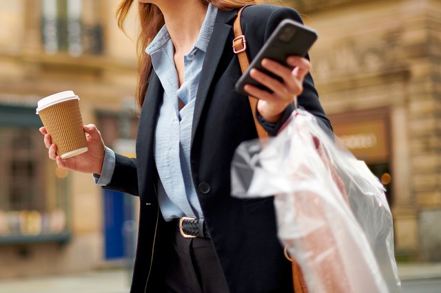 An image of a woman holding a cup of coffee, her phone, and dry cleaning