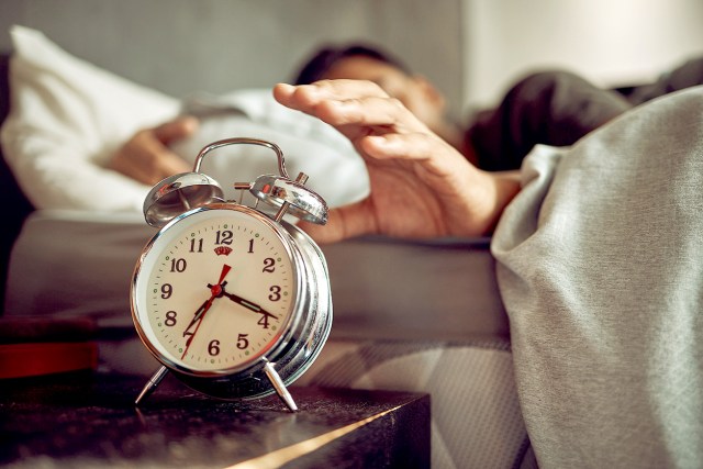 An image of a man in bed reaching for an alarm clock