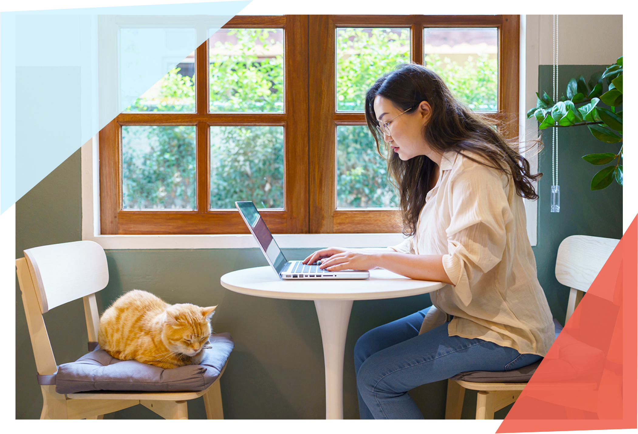 Woman sitting in front of laptop at a table with a cat on the other chair. 