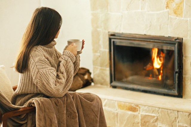 Woman in cozy sweater holding cup of warm tea at fireplace