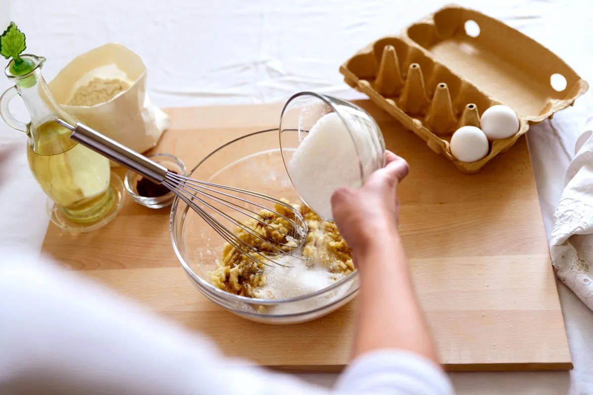 Person pouring sugar into mixing bowl with other baking ingridients 