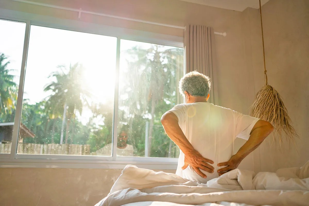 view of a senior man sitting on the bed, holding his hand on back indicating the discomfort 